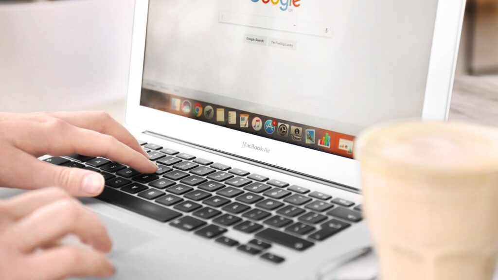 Person using a MacBook Air laptop with Google search engine open on the screen, working at a desk with a coffee in the foreground.
