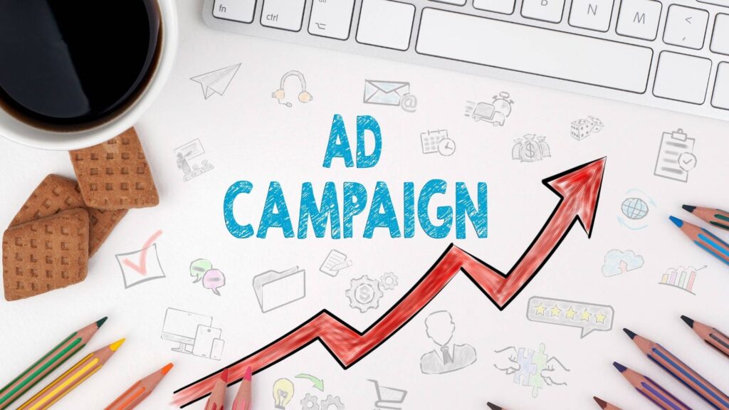 Flat lay image of a workspace showing the words “Ad Campaign” surrounded by colorful marketing icons, a rising red arrow, colored pencils, a keyboard, cookies, and a coffee cup—symbolizing digital advertising growth and creativity.