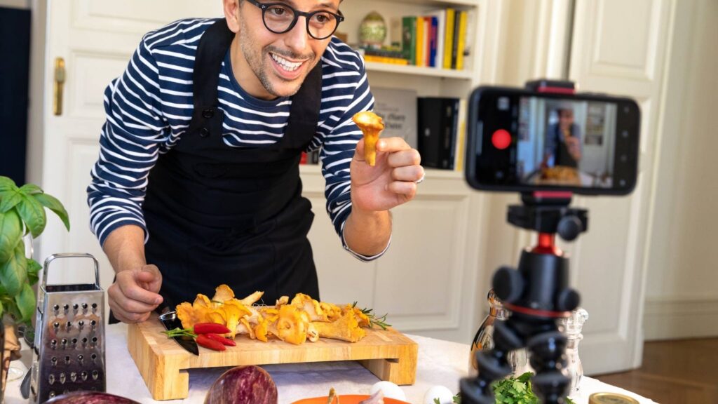A cheerful male content creator films a cooking tutorial using a smartphone mounted on a tripod. He holds up a chanterelle mushroom while explaining ingredients on a kitchen counter filled with vegetables, herbs, and spices—ideal for topics related to food blogging, influencer marketing, or social media video creation.