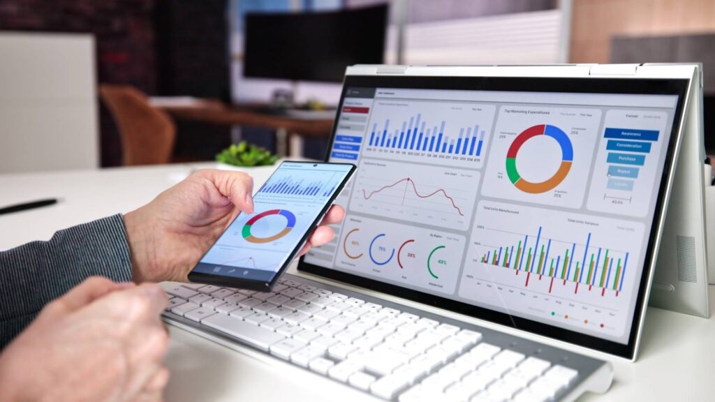 A person analyzing SEO performance metrics on both a smartphone and a laptop screen. The screens display colorful charts and graphs including bar graphs, pie charts, and line graphs representing marketing data, traffic sources, and conversion funnels. A white keyboard and a modern office background are also visible.