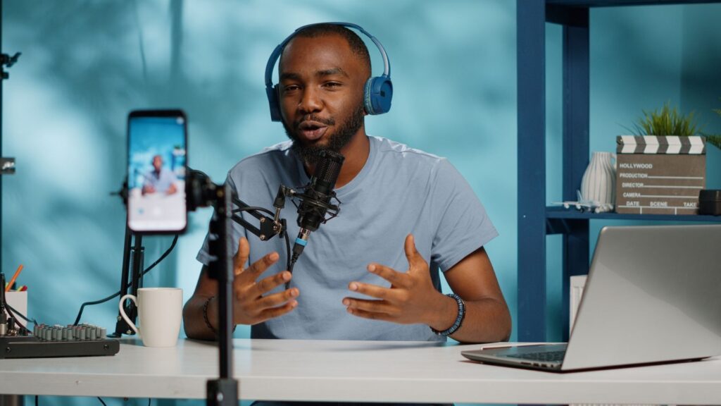 Content creator wearing blue headphones speaking into a microphone while recording a video with a smartphone in a home studio setup. A laptop, coffee mug, and filming equipment are on the desk.